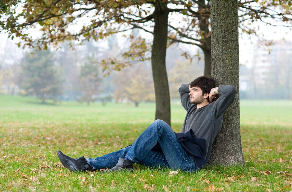 man relaxing, leaning against a tree with his hands behind his head