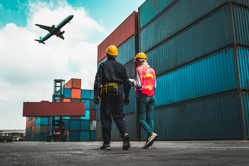 Two transportation workers walking through a container yard with a plane in the sky.