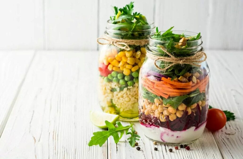Jars filled with lettuce, protein, vegetables, grains, and salad dressings for convenient and healthy lunches.