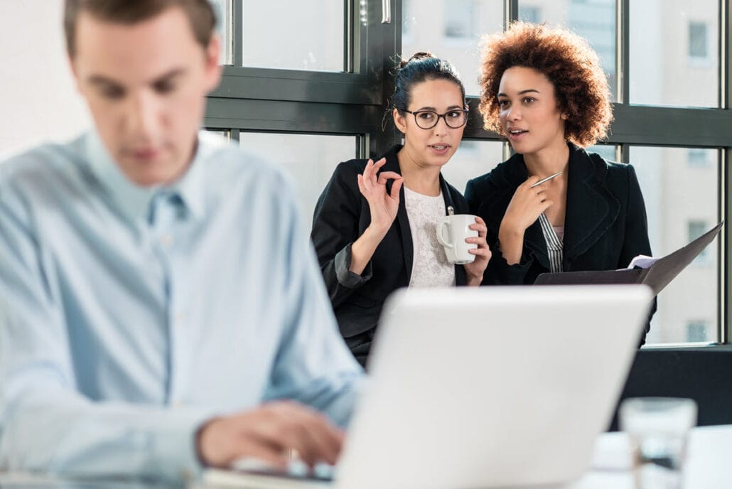 Two women gossiping at work, representing different types of difficult coworkers in a workplace setting.