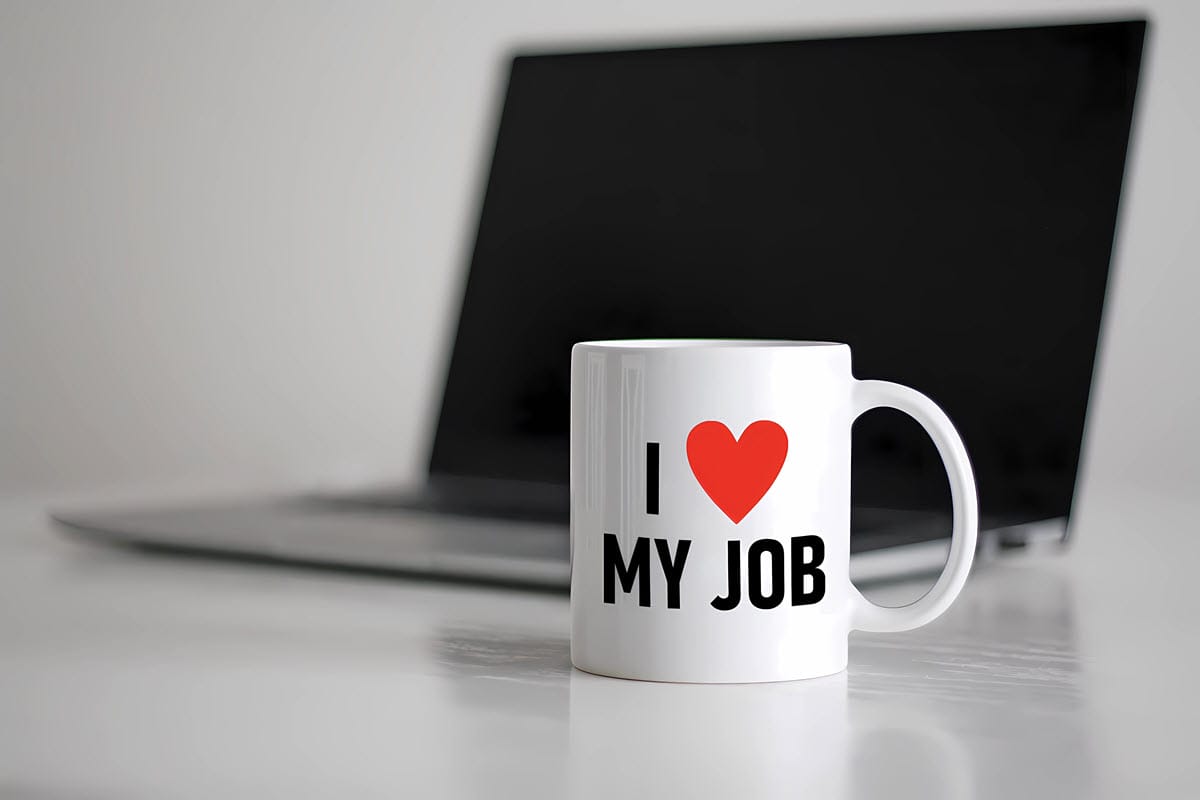 A laptop on a table with a coffee mug in front of it that says I Love My Job