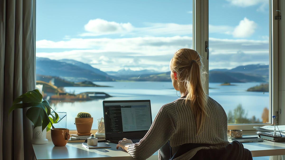 A women working on her laptop in front of a window overlooking a lake and mountainous landscape