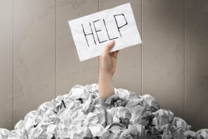 A pile of balled up pieces of paper with a hand reaching through the top holding a sign that says help