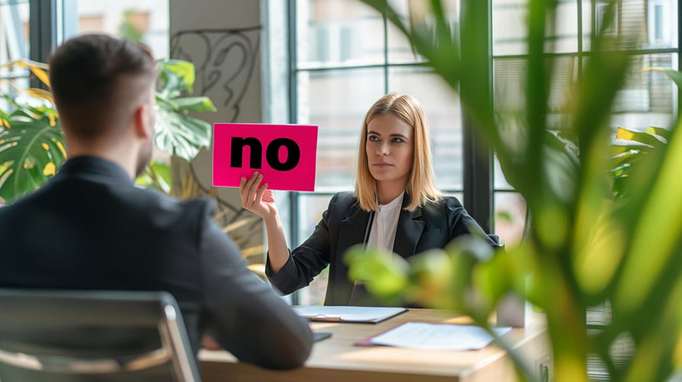 Professional woman at desk holding up a "no" sign to the man across from her, one way to say no at work